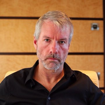 The image depicts an older man wearing a black shirt, sitting comfortably in a chair against a brown wall. The man has a beard and appears engrossed in his thoughts as he looks directly at the camera with an intense expression. His facial hair adds a distinct feature to his appearance. On the screen behind him, there is a chart of Bitcoin's price fluctuations over time, suggesting that the man might be tracking or analyzing this cryptocurrency market. In the post caption provided, it reads: "LATEST: Michael Saylor posted Strategy's Bitcoin chart with ... Bitcoin." This indicates that the image may have been taken recently and that the man is referring to a specific point in time when he evaluated the current value of Bitcoin.