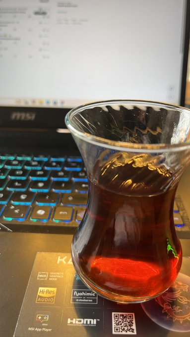 A traditional tulip-shaped glass of dark tea sits on a desk in front of an MSI laptop with a backlit keyboard, displayed on a coaster listing features like "Hi-Res AUDIO," "HDMI," and "MSI App Player" with a QR code—pairing perfectly with a morning "GM" greeting to the web3 community.