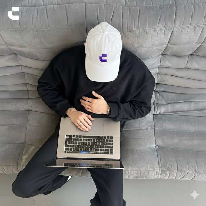 A person wearing a white cap with a purple logo sits on a gray couch with a laptop. This image relates to the "GM" post by potentially representing the start of a day of work or productivity.