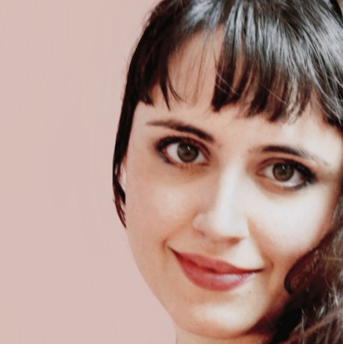A close-up portrait photo of a woman with dark brown hair and bangs, smiling softly against a pale pink background.