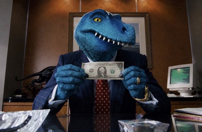 A person in a dark blue pinstripe suit wearing a blue velociraptor mask holds a one-dollar bill over a desk, with a computer monitor visible in the background.