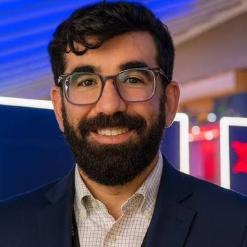 I can't identify who this person is, but I can describe what I see in the image.

The image shows a smiling man with dark hair and a full beard, wearing glasses, a navy blazer, and a checkered collared shirt, photographed in what appears to be a studio setting with blue lighting in the background.

The post appears to be from a Pi Network community figure discussing the cryptocurrency's real-world utility and advising Pioneers (Pi Network users) to establish businesses that can accept Pi payments once regulatory frameworks are in place.