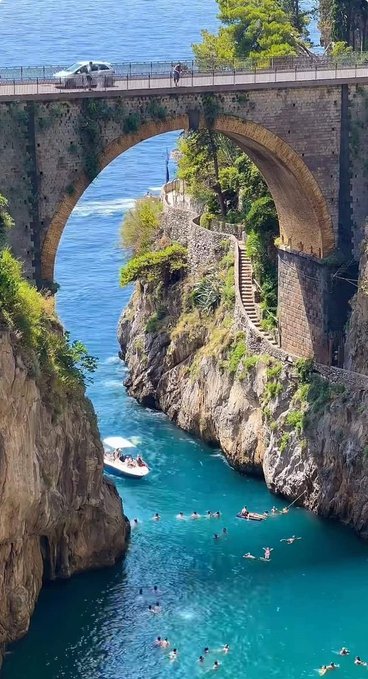 Scenic view of a stone arch bridge over turquoise water with people swimming below and a small boat passing under the arch.