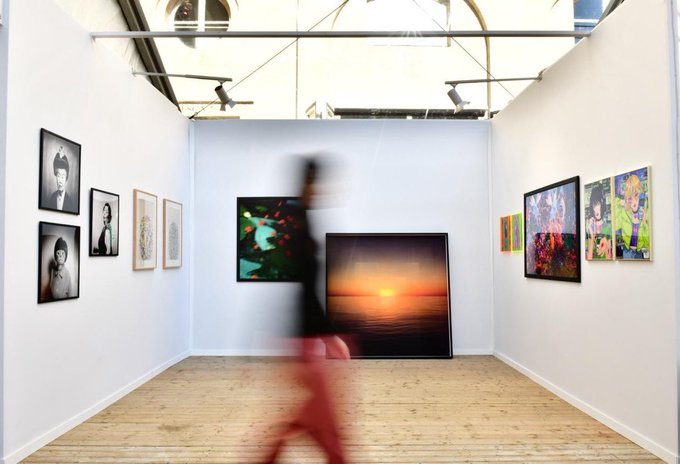 A blurred figure in red pants walks through a white-walled art gallery, where various framed portraits and colorful paintings are displayed on the walls.