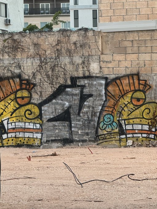 A weathered brick and concrete wall covered in colorful graffiti depicting stylized cartoonish faces with large eyes and teeth, flanked by apartment buildings visible above.