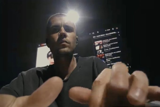A man with short hair and an earring reaches toward the camera with his hand in the foreground, while a computer screen behind him displays a social media profile page with visible post thumbnails and follower information. The low-light, close-up composition humorously conveys his eager anticipation to check whether others have seen his post after logging into his laptop.