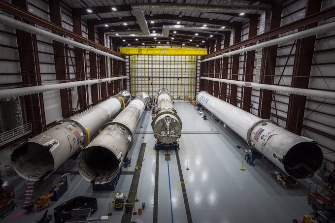 A view inside SpaceX's integration facility showing three Falcon 9 first-stage boosters lined up horizontally, with the American flag visible on the rightmost rocket and the "SPACEX" logo on the center booster, awaiting processing. The mention of #SpaceXcoin plays on the visual of these reusable boosters, highlighting the company's connection to spaceflight.