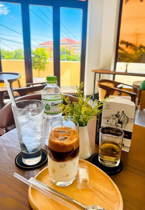 A café table with an iced coffee, a glass of water, a bottle of Dasani water, a small vase of yellow flowers, and a glass of brown liquid, set against a window view of buildings and palm trees.