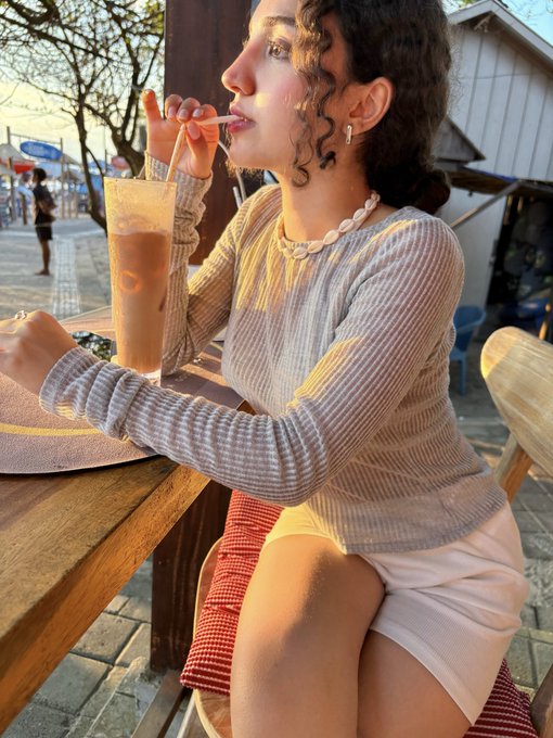 A young woman with curly dark hair drinking an iced beverage through a straw at an outdoor café, wearing a grey ribbed long-sleeve top and a shell necklace, bathed in warm sunset light.