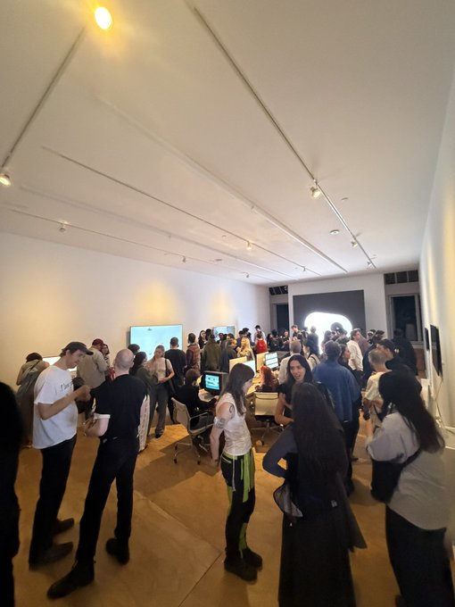 A crowd of people gathers in a bright, white-walled gallery space, with a woman in a white top and green/black pants standing in the foreground looking at the scene.