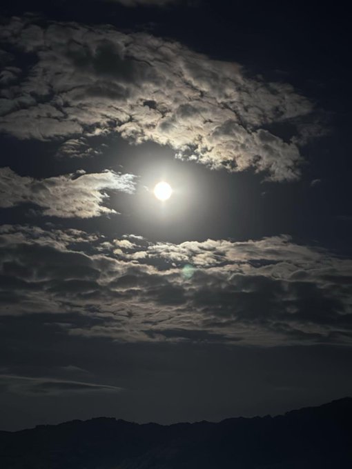 Bright full moon shining through scattered clouds in a dark night sky, with the silhouette of a mountain ridge at the bottom of the frame.