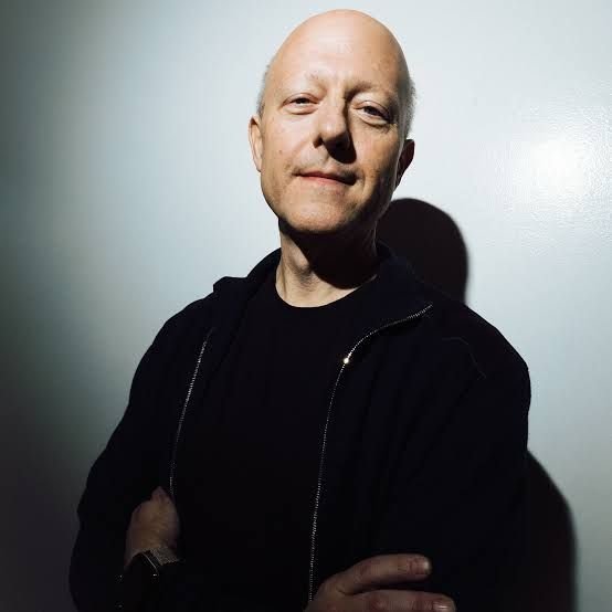 A bald man in a black zip-up jacket stands against a gray wall with his arms crossed, offering a slight smile in a moody, dimly lit portrait. The image pairs with the post's call for him to "press the MAINNET button," visually portraying the individual being urged to launch the network.