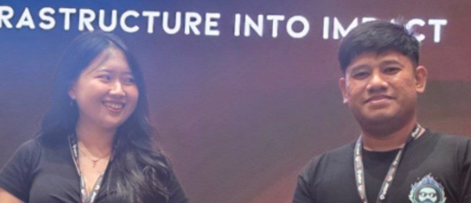 Two people pose for a photo in front of a dark backdrop with partial text reading 'RASTRUCTURE INTO IMPACT'. The woman on the left wears a black top and lanyard, while the man on the right wears a dark shirt with a logo.
