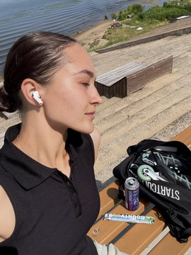 A young woman wearing white earbuds sits outdoors by a river, eyes closed, with a backpack labeled 'NATION' and an energy drink beside her.