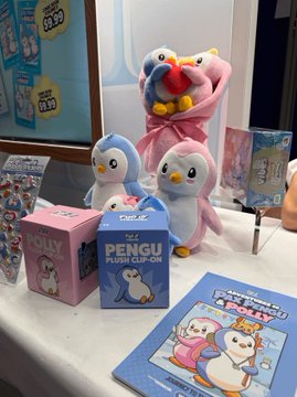 A display of penguin-themed merchandise including plush toys, a 'Pengu Plush Clip-On' box, and various packaged products on a store shelf.
