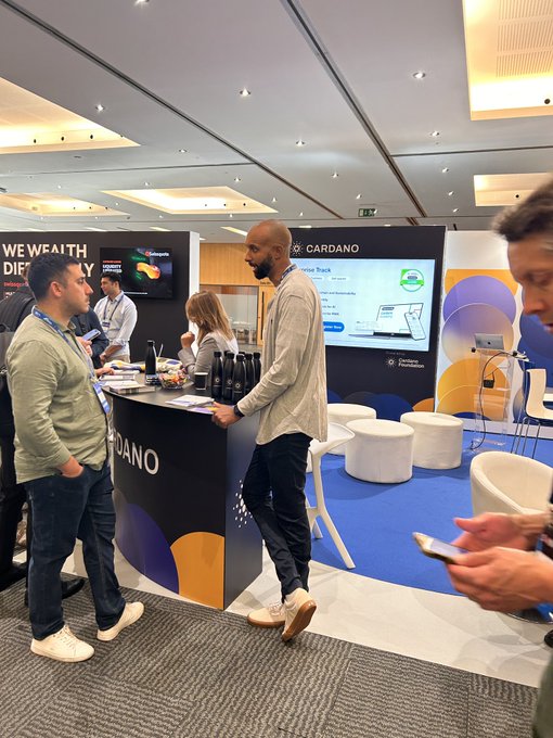 A man in a gray sweater and cap leans on a counter at a CARDANO booth at what appears to be a conference, while others stand around him and a person in the foreground looks at their phone.