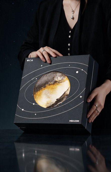 A woman in a black blouse holds up a black "MSX.COM" gift box featuring a gold moon design surrounded by orbital rings and stars, with the box's reflection visible on the glossy surface below. The celestial moon imagery on the packaging directly ties to the Mid-Autumn Festival celebration theme of the post.