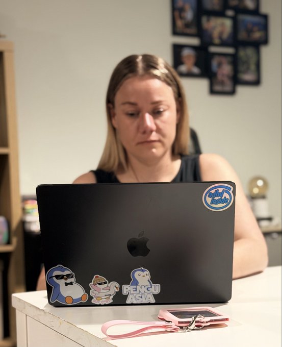 A woman works at a desk behind a dark MacBook laptop decorated with cartoon penguin and character stickers, including one reading "PENGU ASIA."