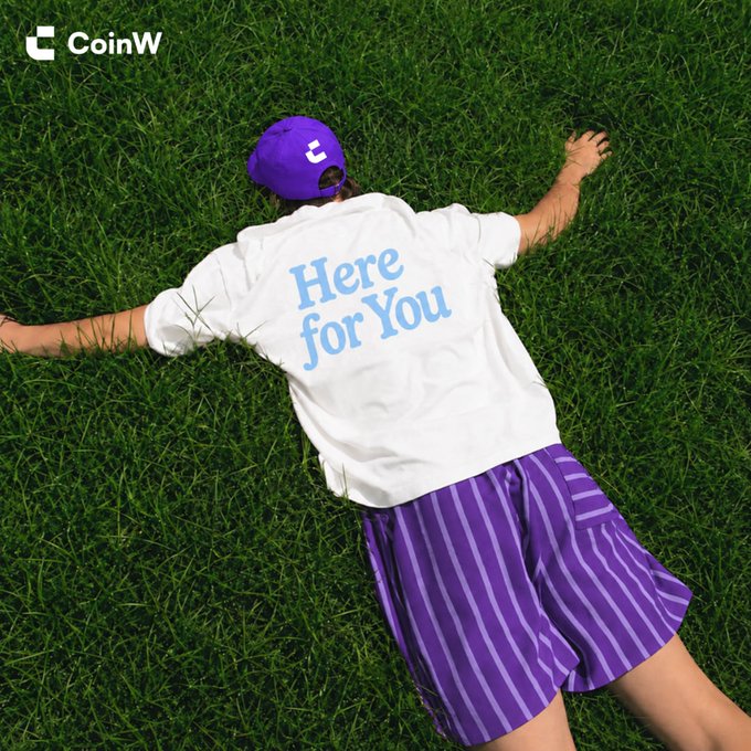 A person lies face-down on lush green grass, wearing a purple "CoinW" cap, a white T-shirt with "Here for You" printed in light blue, and purple striped shorts, visually answering the post's "Guess who's always here for you?" by embodying the CoinW brand's supportive message.