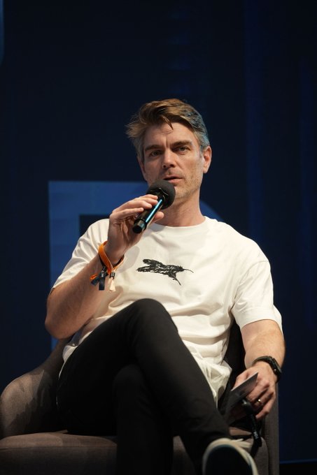 A man with tousled blonde hair sits on a dark armchair, holding a microphone to his mouth as he speaks on a stage with deep blue lighting. He wears a white t-shirt featuring a black fish graphic, an orange wristband, and black pants, appearing engaged in a panel discussion about Hyperliquid's institutional challenges.
