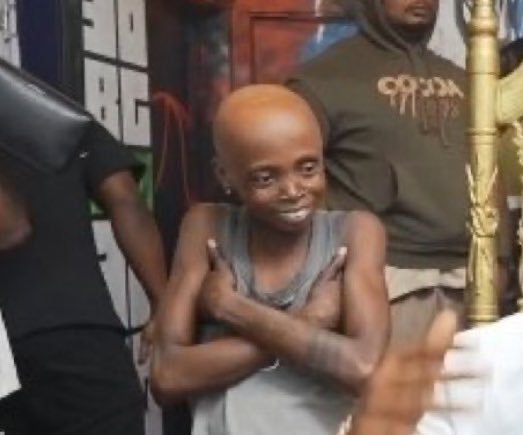 A young bald child smiles with arms crossed while standing in front of an adult wearing a 'G-Unit' hoodie and another person in black.