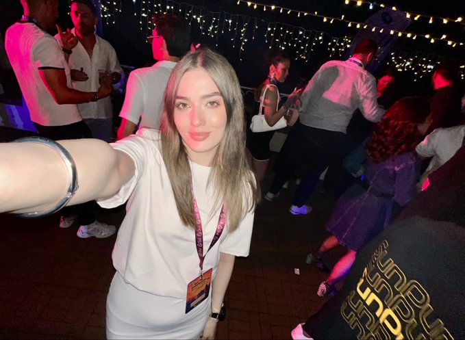 The image features a young woman wearing a white shirt, standing next to a brick wall with other people around her. She has taken a selfie using her phone while at an event or gathering. The background of the photo shows multiple people enjoying themselves, indicating that they are attending a party or social event together.