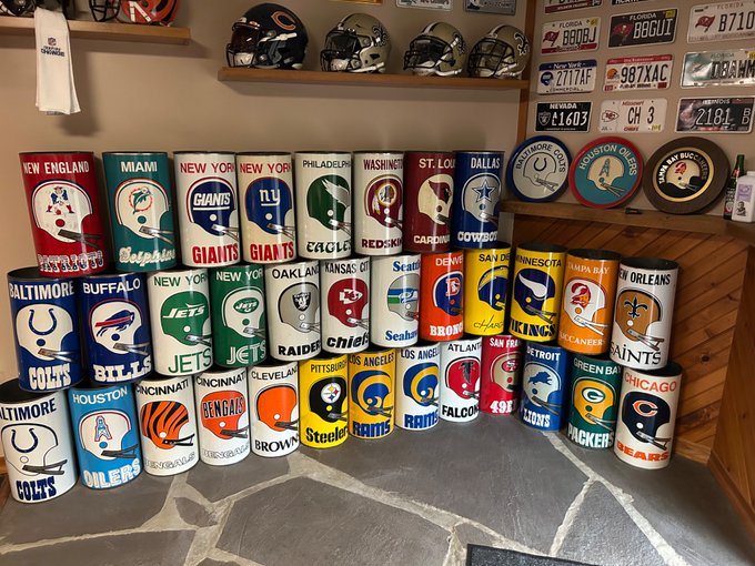 Display collection of NFL team can coolers/koozies arranged in rows featuring teams like Patriots, Dolphins, Giants, Jets, Cowboys, Eagles, Raiders, Chiefs, 49ers, and Packers, with football helmets and license plates visible on shelves above.