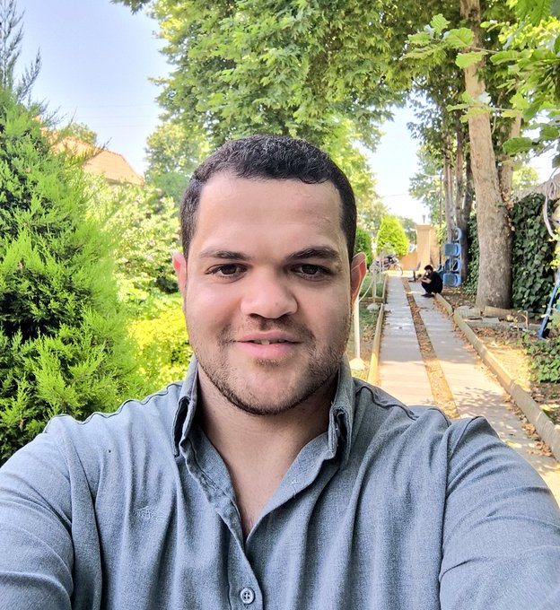 A selfie of a smiling man with a short beard wearing a blue denim shirt, standing outdoors on a sunny day with green trees and a pathway in the background.