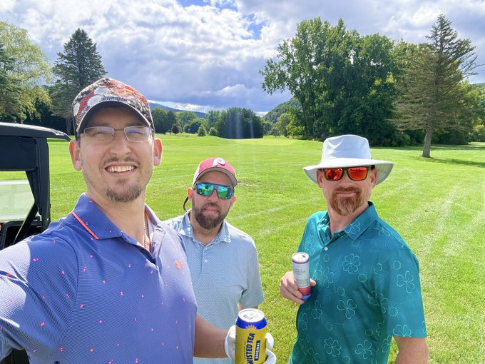 Three smiling men on a golf course posing for a photo, wearing colorful hats and sunglasses, with one holding a Twisted Tea can.