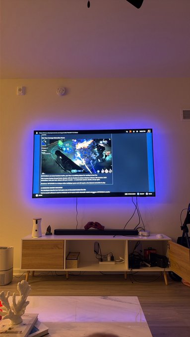A living room scene featuring a large TV displaying a video game with text, mounted on a wall with purple backlighting above a wooden TV stand holding a game console, controller, and other electronics.