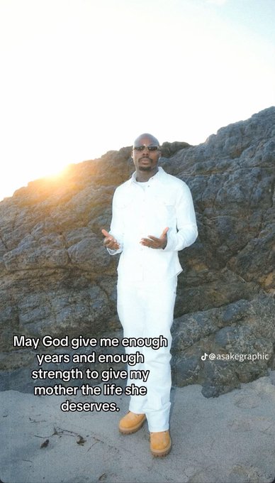 A man dressed in an all-white outfit stands on a rocky beach at sunset with his hands raised in a prayerful gesture, accompanied by the text "May God give me enough years and enough strength to give my mother the life she deserves," shared by @asakegraphic. The image conveys a heartfelt morning prayer ("Gm Guys") expressing gratitude and a wish to bless his mother.
