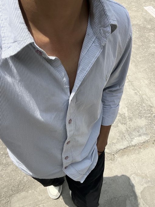 A close-up downward view of a person wearing an unbuttoned light blue striped shirt and black pants, standing on cracked concrete pavement with white sneakers visible.