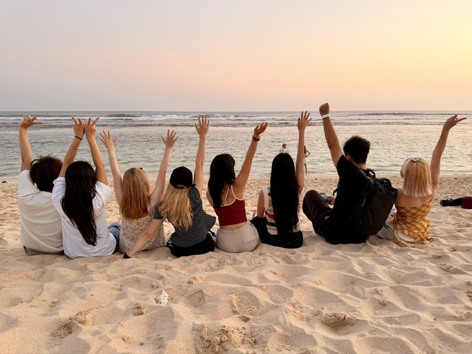 A group of people sitting on a beach at sunset with their hands raised in the air facing the ocean.