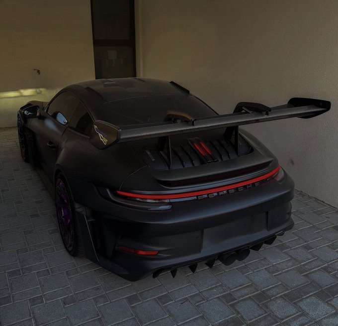 A matte black Porsche 911 with a large rear wing, aggressive aerodynamic bodywork, purple-accented wheels, and a red LED taillight bar, parked on gray brick pavers under covered lighting.