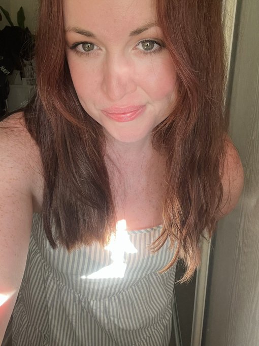 A selfie of a red-haired woman with freckles wearing a striped top, standing indoors with a plant visible in the background.