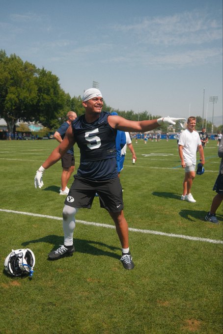 A young football player wearing a navy 'S' jersey, black shorts, and a bandana smiles and gestures on a grass practice field during a sunny day.