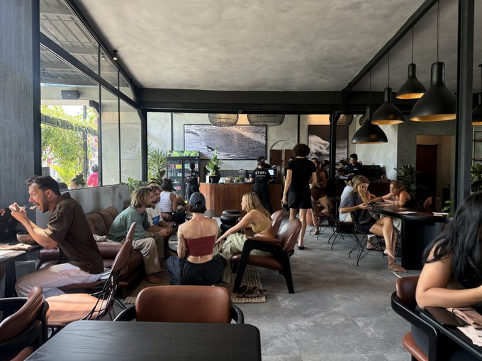 A busy, modern cafe interior with industrial decor, pendant lights, and brown leather seating, where several customers are sitting at tables or waiting in line.