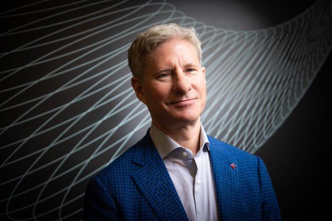 Chris Larsen, co-founder of Ripple, smiles in a professional headshot wearing a blue blazer, with abstract flowing line patterns in the background—visually representing his advocacy for advancing crypto regulatory frameworks like the CLARITY Act.