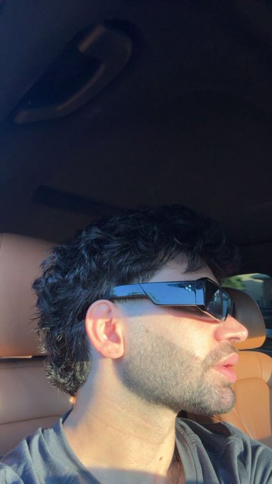 A side profile selfie of a man with curly dark hair wearing modern wraparound black sunglasses, sitting in what appears to be a vehicle with tan leather seats.
