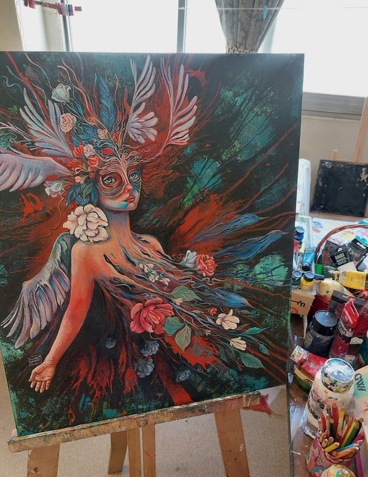 A vibrant expressionist painting on an easel depicts a winged woman with floral elements, surrounded by paint tubes and brushes in an artist's studio.
