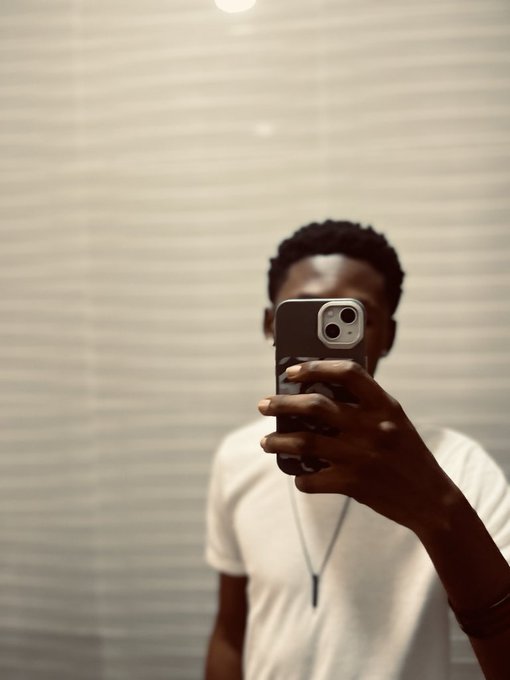 A man in a white t-shirt and silver necklace takes a mirror selfie with his smartphone in a room with closed blinds.