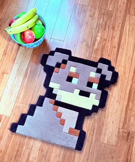 A pixel-art style rug depicting a bear or similar animal character, placed on a wooden floor next to a bowl of fruit containing bananas, apples, and a lime.