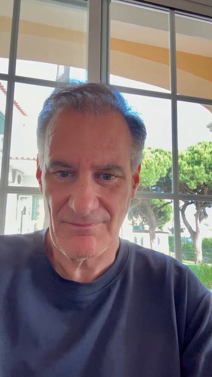 The image features a man wearing a blue shirt, sitting at a table with his head tilted back towards the camera. The background of the photo shows an open window that provides natural light into the room. There are also some trees visible outside the window. The image is related to Bitcoin because it contains text about Bitcoin and its value being drained by a company called Saylor.