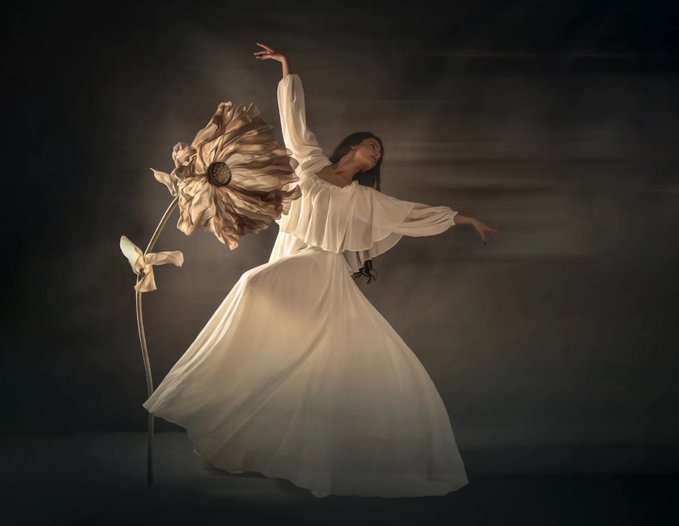 A woman in a flowing white dress poses gracefully beside a large dried sunflower in a dimly lit, atmospheric studio setting.