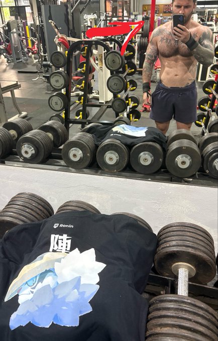 A shirtless, tattooed man takes a mirror selfie with his phone in a gym, standing behind a large rack of heavy dumbbells, while an anime-themed 'Ronin' t-shirt rests on weights in the foreground.
