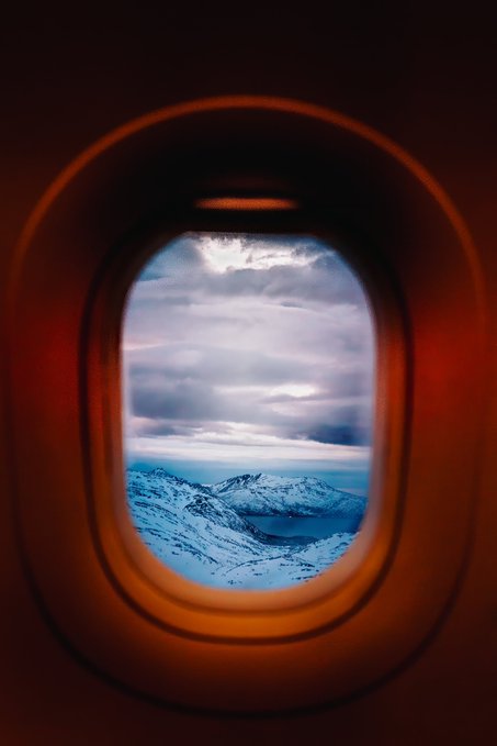 A view through an airplane window showing snow-covered mountains and a cloudy sky below, with a warm orange glow on the interior window frame.