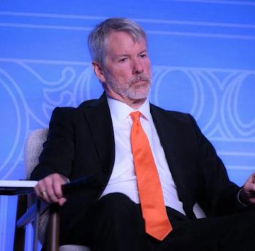 The image shows a man wearing a suit and tie sitting in a chair with his hands resting on the armrest, looking off into the distance. The background features a blue wall adorned with white designs. There are no other people or objects visible in the image.
