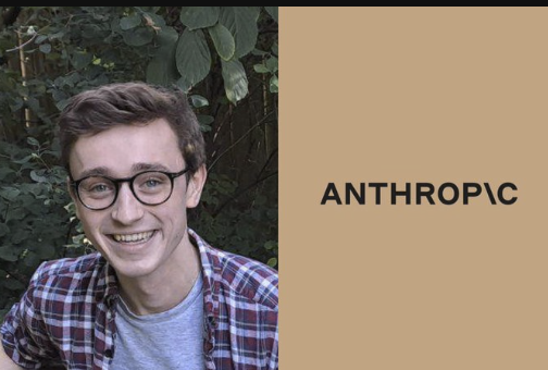 A young man with glasses and a plaid shirt smiles outdoors against green foliage, next to a tan panel displaying the 'ANTHROPIC' logo.