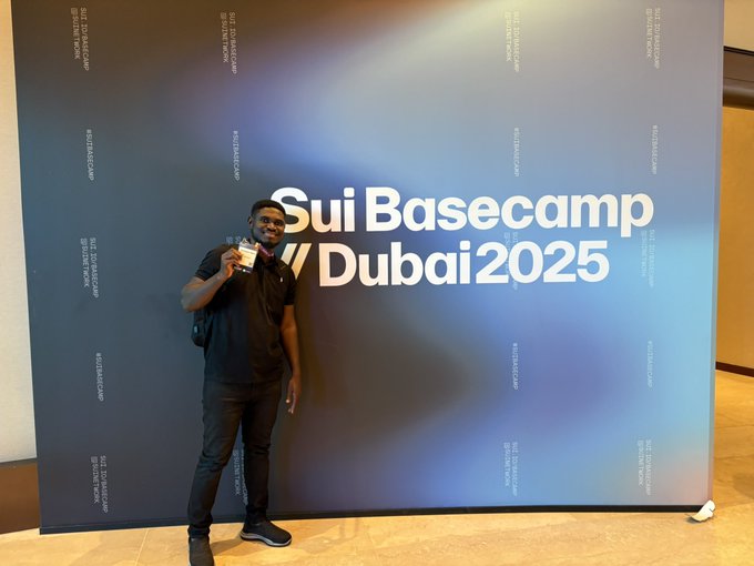 A man wearing sunglasses and a black shirt stands in front of a large blue backdrop reading 'Sui Basecamp Dubai 2025', holding a coffee cup.
