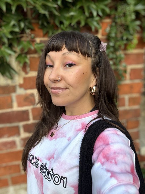 A young woman with dark hair, blunt bangs, pink eyeshadow, a nose ring, and gold hoop earrings stands against a brick wall with greenery, wearing a pink and white tie-dye sweatshirt with 'Love sickness' printed on it.